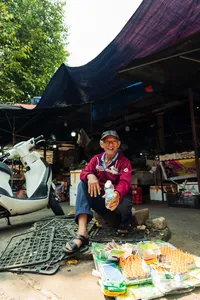 会安市场摊位一景：一位亲切的摊主坐在遮棚下，手里拿着饮料瓶，身旁是包装商品和一辆摩托车