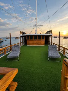 Sunset deck on a wooden phinisi with green carpet, padded loungers, and anchored masts overlooking the harbour in Labuan Bajo