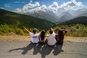 Travellers sitting on a roadside viewpoint above Shangri-La, arms raised toward snowy peaks during a 6-day Loop with a Tibetan guide