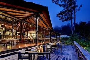 Borneo Rainforest Lodge veranda with dining tables overlooking Danum Valley rainforest at dusk, framed by tall trees and soft lights