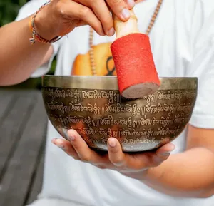 Close view of hands ringing an embossed singing bowl with a red mallet during sound healing on an outdoor mat in Canggu