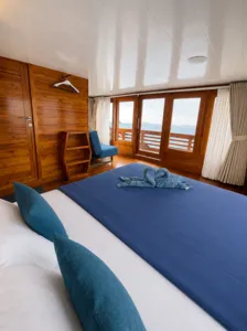 La Famille cabin bedroom on a wooden phinisi with a sea-view window, neatly made beds and towels for a Komodo cruise Labuan Bajo