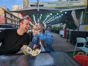 Travellers tasting durian at a busy Geylang night market stall in Singapore, smiling with wedges under hanging lights