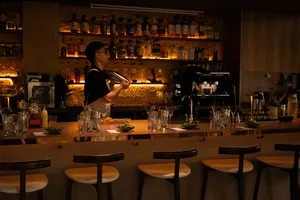 Bar koktail bercahaya hangat di Singapura dengan bartender di belakang meja bar, botol-botol di rak, dan gelas-gelas siap diisi