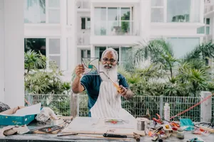 Elderly glass artist shaping a curved stained-glass panel at a table in a bright garden, Singapore—workshops in Singapore