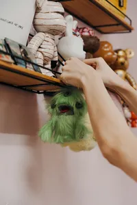 Hand placing a green ugly-cute plushie keychain on a display shelf of stuffed characters at Avant-Garde Art Space workshops in Singapore