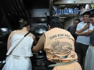 Back view of visitors in 'Tang Kay Kee' aprons standing at a busy hawker kitchen with large pots in Chinatown Singapore