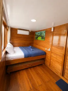 Corner wooden phinisi cabin with queen bed, curtain window and wall artwork on a shared Komodo cruise from Labuan Bajo