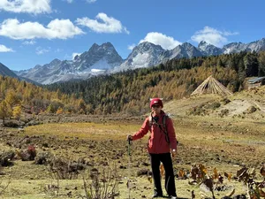 Hiker in a red jacket stands on a grassy trail with distant snow peaks and forested slopes in Shangri-La during the trek