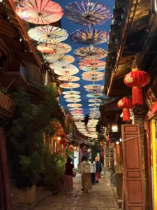 Lantern-lit lane in Lijiang’s old town with colourful paper umbrellas overhead as travellers stroll at dusk