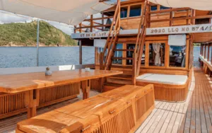 Wooden sun deck with built-in benches and shaded loungers on a deluxe phinisi, cruising along the coast near Labuan Bajo