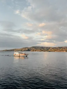 Sunset cruise view: a Deluxe phinisi gliding across calm water beside low islands as the sky darkens on the Komodo route, Komodo Island