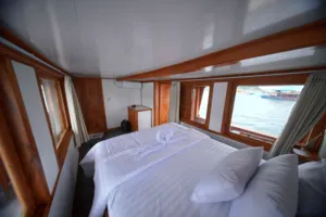 Cozy wooden boat cabin with a made double bed and wide windows looking out to the harbour water in Labuan Bajo