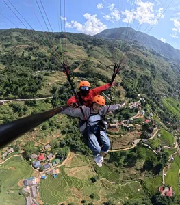 Tandem paragliding with an orange-helmeted pilot and passenger soaring above green mountain slopes and village farmland in Sapa