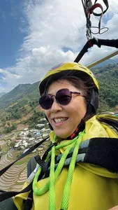 Smiling paragliding passenger in a yellow helmet and sunglasses looks out over green terraces and a river valley under blue sky in Sapa