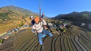 Helmeted paraglider flying above patchwork terraced fields and rooftops, legs lifted, with Sapa Paragliding over Sapa