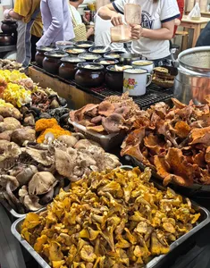 Trays of cooked meats and mushrooms at a Lijiang market stall, with sizzling pots behind and diners with cups