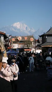 Crowded narrow market street in Lijiang old town with traditional shops, Chinese signboards, and snow-dusted mountains in view