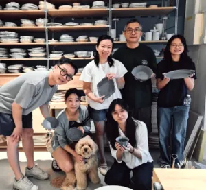 Ina Atelier pottery studio group holding handmade ceramic plates, with shelves of bisqueware in the background during workshops in Singapore
