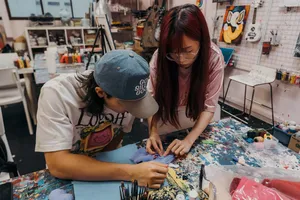 Two participants stitching an ugly-cute plush keychain on a shared table in a Singapore art studio, surrounded by tools