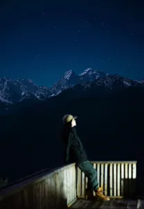 Person in a dark jacket stands on a wooden viewing platform under a starry night sky over mountains in Shangri-La