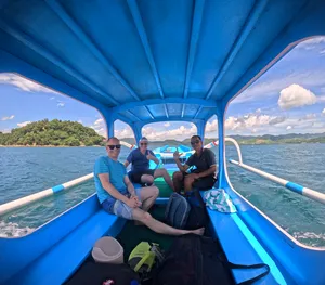 Para penumpang tersenyum dan melambai di atas bot bermotor biru dalam perjalanan ke Kepulauan Gili untuk aktiviti snorkeling persendirian berhampiran Lombok