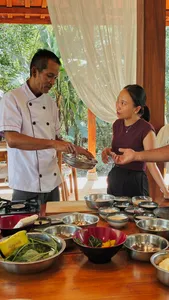 Chef demonstrates a vegan Manggarai cooking step to a participant at a table of ingredients during a class in Labuan Bajo