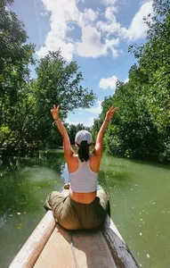 Woman sitting on a boat deck with arms raised above calm green water and thick trees during a mangrove visit in Labuan Bajo
