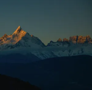 Sunlit snow-capped mountain peaks above dark ridgelines in Shangri-La at dusk, with a dramatic clear sky fading to blue
