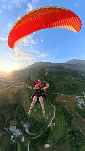 A paraglider duo flies beneath a bright red canopy over winding roads and villages during golden hour in Sapa