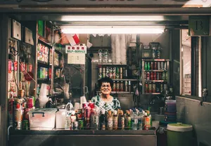 Chef in a Singapore hawker stall serving from behind a counter lined with jars and tins, with a “self-service” sign