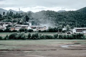 Pemandangan desa dari retret ramah lingkungan Linden Centre di Shaxi, Dali, dengan atap-atap halaman, hamparan ladang, dan pegunungan berkabut di balik pepohonan