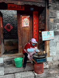 Street food cook tending a small stove on a stone step in Lijiang Old Town, beside hanging Chinese signs and a wooden door