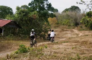 Cyclists ride on a sandy track through Langkawi countryside, passing a small roadside house and dense greenery