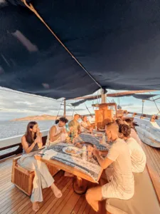 Shared dining on the deck of a deluxe phinisi in Labuan Bajo, with travellers enjoying drinks together, Komodo Island