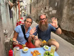 Smiling travellers waving from a street-feast table in a narrow Hanoi alley with bowls of food and drinks