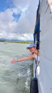Seorang pengembara tersenyum membongkok di atas badan perahu layar putih di perairan hijau yang beralun semasa pelayaran di bawah langit biru, Labuan Bajo