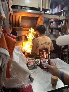 Cook tending a flaming wok at a Singapore hawker stall kitchen, while diners watch and take a photo with a phone