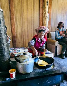 Lijiang village kitchen where a local woman cooks by a stove, with another person seated nearby on a day meal
