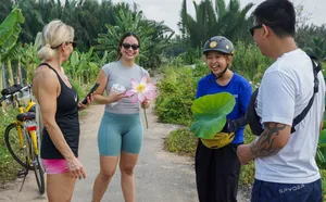Cyclists on a narrow dirt track in lush greenery, one holding a pink lotus, during a bike tour of Mekong Island near Ho Chi Minh City
