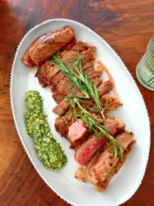Hirisan daging dengan rosemary di atas pinggan putih bergaya antik, dihidangkan bersama sos herba hijau di sisi