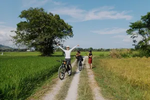 Cyclists pedal along a narrow countryside lane in Langkawi, passing bright green rice fields under a blue sky
