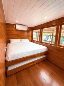 Wooden phinisi cabin with a double berth and side window inside a 3D2N shared Komodo cruise, Komodo Island