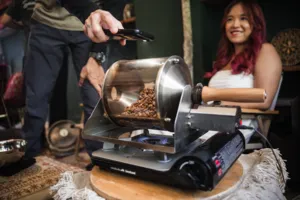 Beans being tipped into an electric coffee roaster as a woman smiles in the background during hands-on coffee roasting workshops in Singapore