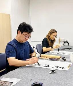 Chinese calligraphy workshop in Singapore with two learners writing on rice paper at tables, brushes and ink ready