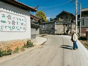 Traveller walking along a narrow lane in a Lijiang village, with traditional walls and distant rooftops on the Yi & Naxi trek