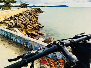 Bike handlebars on a Langkawi coastal route beside a rocky breakwater, with calm sea stretching to distant islands