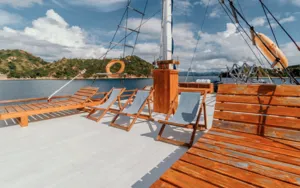 Sunlit wooden deck with loungers and sail rigging on a deluxe phinisi cruise, departing over Labuan Bajo