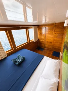 La Famille wooden phinisi cabin with two beds made up, large sea-facing windows and calm ocean outside on a Komodo cruise Labuan Bajo